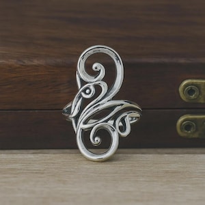 May include: A silver ring featuring an elongated, openwork design composed of polished, swirling metal scrolls and curved lines. The metal surface has a bright, reflective finish with darkened recessed areas that emphasize the intricate, flowing pattern.