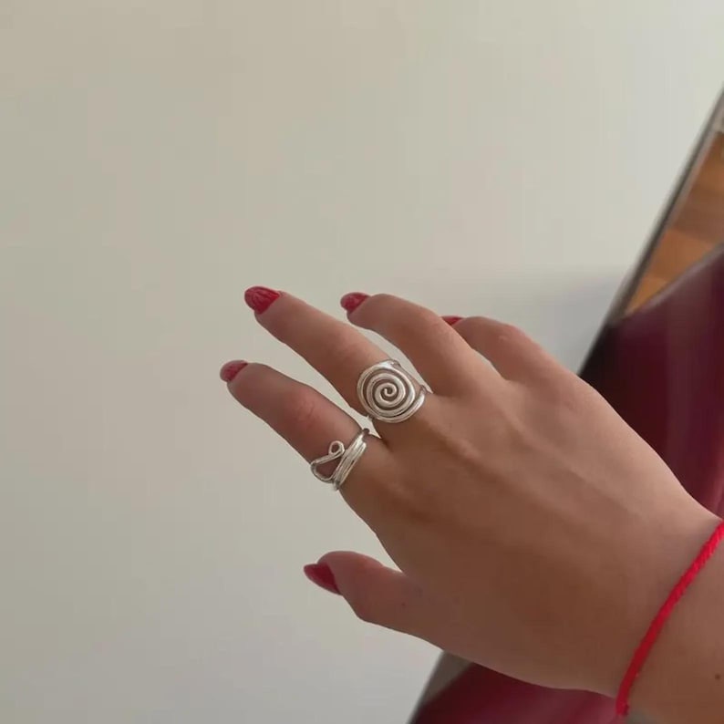 May include: Two silver rings are displayed on a hand with red nail polish. One ring features a spiral design, while the other has a curved, open shape. A red bracelet is visible on the wrist.