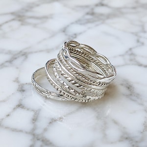 May include: A stack of silver rings with various textures, including braided, beaded, and etched designs. The rings are displayed on a white and gray marble surface, showcasing their intricate details and reflective qualities. The rings are a jewelry item.