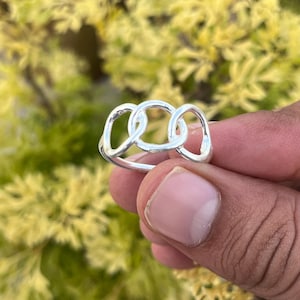 Könnte beinhalten: Ein silberner Ring mit einem verbundenen Design aus drei ineinandergreifenden ovalen Formen. Der Ring besteht aus glänzendem Metall und wird vor einem verschwommenen grün-gelben Hintergrund gehalten.