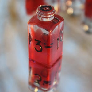 May include: A clear, square-shaped bottle filled with a vibrant red liquid. Black numbers and symbols are painted on the sides. The bottle is reflected in the surface below. The top of the bottle has a black number 4.