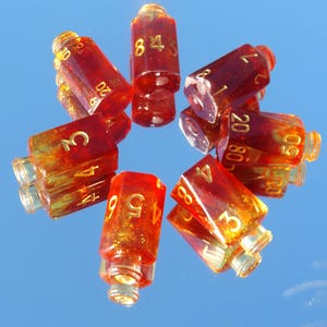 May include: A collection of transparent, red-orange dice with gold numbering. The dice are arranged in a circular pattern, reflecting on a mirrored surface. The numbers are clearly visible, indicating their use for tabletop games.