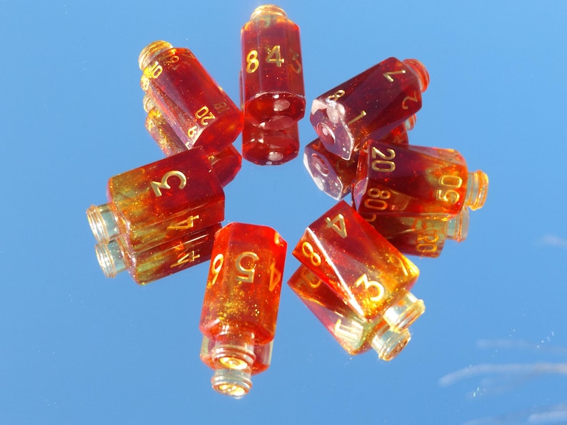 May include: A collection of transparent, red-orange dice with gold numbering. The dice are arranged in a circular pattern, reflecting on a mirrored surface. The numbers are clearly visible, indicating their use for tabletop games.
