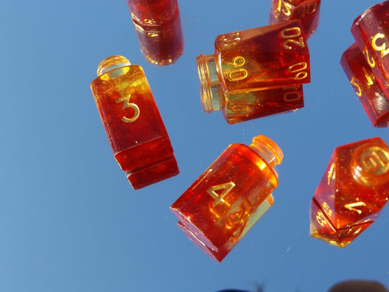 May include: Several amber-colored dice with gold numbering are scattered against a blue background. The dice are various shapes, including rectangular and multi-sided. The numbers 3, 4, 10, 15, 20, 60, and 90 are visible.