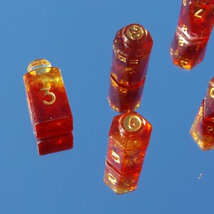 May include: Several red and gold resin dice with gold numbering are scattered against a blue background. The dice are various shapes, including a rectangular die with the number 3, and a 20-sided die. The dice have a glittery appearance.