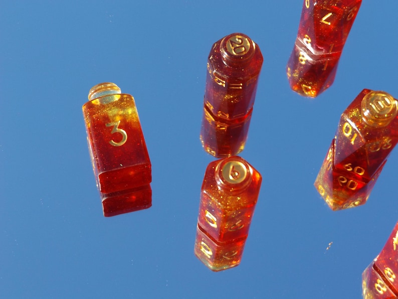 May include: Several red and gold resin dice with gold numbering are scattered against a blue background. The dice are various shapes, including a rectangular die with the number 3, and a 20-sided die. The dice have a glittery appearance.