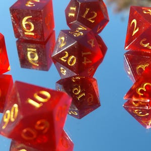 May include: A collection of translucent red polyhedral dice with gold numbering. The dice are various shapes, including six-sided, eight-sided, ten-sided, twelve-sided, and twenty-sided. The dice are scattered on a reflective surface.