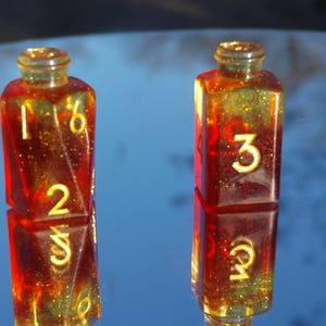 May include: Two resin dice in a bottle shape, with a red and gold glitter effect. The dice have yellow numbers, with one showing 1, 2, and 6, and the other showing 3, 4, and 5. The dice are reflected on a shiny surface.