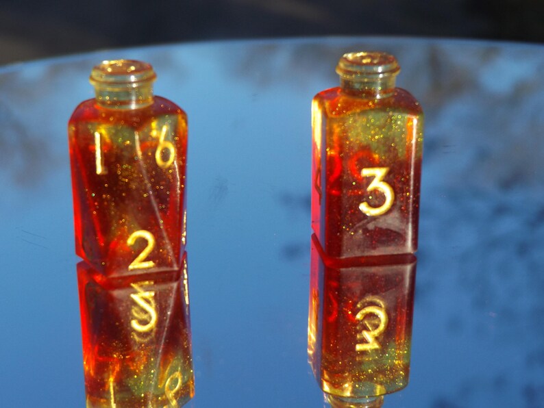 May include: Two resin dice in a bottle shape, with a red and gold glitter effect. The dice have yellow numbers, with one showing 1, 2, and 6, and the other showing 3, 4, and 5. The dice are reflected on a shiny surface.