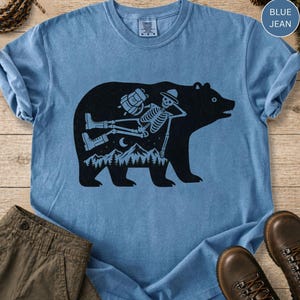 Op de afbeelding: Een blauw spijkerkleurig t-shirt met een grote zwarte afbeelding van een berensilhouet. Binnenin de berenvorm ligt een skelet met een hoed en laarzen, met een rugzak, boven een landschap van bergen, bomen en een halve maan.