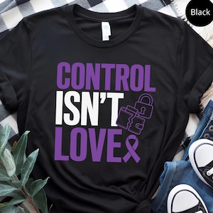 Könnte beinhalten: Ein schwarzes Kurzarm-T-Shirt mit dem violett-weißen Schriftzug "CONTROL ISN'T LOVE" auf der Vorderseite. Das Design enthält eine Grafik eines zerbrochenen Vorhängeschlosses und eine violette Schleife, die in den Text integriert ist.
