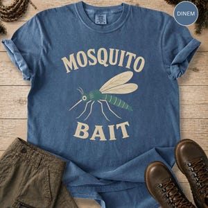 May include: A blue short-sleeve t-shirt featuring a graphic of a green mosquito with beige wings. The text "MOSQUITO" is printed in beige above the insect, and the word "BAIT" is printed in beige below it. The shirt has a crew neckline.