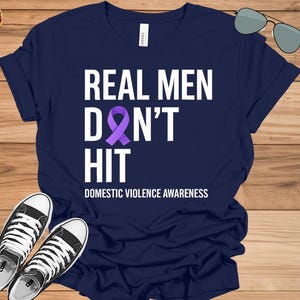 Könnte beinhalten: Ein marineblaues T-Shirt mit dem Text "REAL MEN DON'T HIT" in Weiß, einem lila Band und "DOMESTIC VIOLENCE AWARENESS" in Weiß. Schwarz-weiße Turnschuhe sind unten.