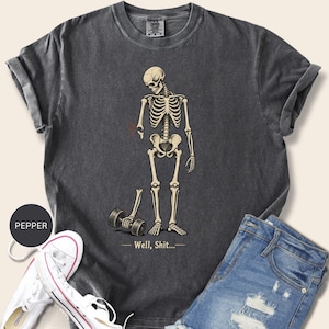 Könnte beinhalten: Ein anthrazitfarbenes T-Shirt mit einer Grafik eines Skeletts, dessen abgetrennter Unterarm eine Hantel hält. Das Skelett ist mit einem gebrochenen Arm dargestellt und der Text darunter lautet "Well, Shit...". Das T-Shirt hat einen Rundhalsausschnitt.
