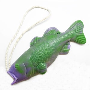 May include: A green and purple fish-shaped soap with a white rope attached. The fish has a textured surface and is designed with a detailed scale pattern. The soap is a novelty item.