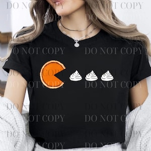 以下が含まれることがあります： 黒いTシャツ。パイをテーマにしたパックマンのデザイン。パックマンのキャラクターはパイのスライスで表現され、3つの白い幽霊のような形があります。シャツはクラシックなクルーネックと半袖です。