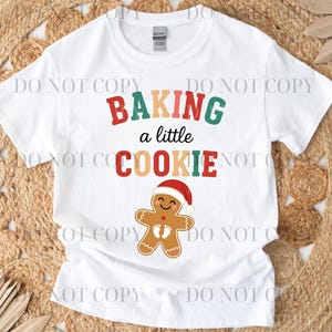 May include: White t-shirt with the text "BAKING a little COOKIE" in red, green, and tan colors. A smiling gingerbread man wearing a Santa hat is below the text. The shirt is made of a soft material.