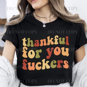 Puede incluir: Camiseta negra con la frase "thankful for you fuckers" en letras de estilo retro. El texto es en tonos de verde, naranja y amarillo. La camiseta tiene cuello redondo y mangas cortas.