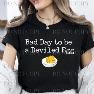 May include: Black t-shirt with the text "Bad Day to be a Deviled Egg" in white. Below the text is an illustration of a deviled egg. The shirt is a casual style.