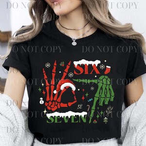 Puede incluir: Camiseta negra con un diseño navideño. El diseño presenta una mano esquelética roja formando el número siete, una mano esquelética verde formando el número seis, y las palabras "SIX" y "SEVEN". Incluye copos de nieve, un gorro de Papá Noel y un bastón de caramelo.