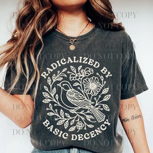 以下が含まれることがあります： 白のグラフィックが入った濃いグレーのTシャツ。グラフィックは鳥、花、そして「RADICALIZED BY BASIC DECENCY」というテキストを円形デザインで表示しています。Tシャツは半袖で、クルーネックです。