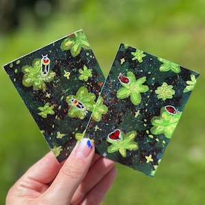 May include: Two rectangular cards featuring a dark background with a pattern of bright green flowers and small red and white insect illustrations. The surface of the cards has a speckled, multi-colored texture with tiny dots of paint throughout.