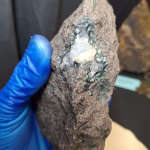 May include: A rough, dark brown rock with a cavity revealing a light-colored crystal formation. The edges of the cavity are lined with small, dark green mineral deposits. The rock is held in a blue gloved hand, suggesting a geological specimen.