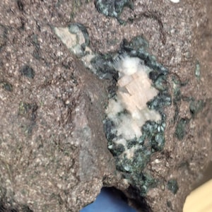 May include: Close-up of a rough, reddish-brown rock with embedded minerals. Patches of dark green and white minerals are visible, along with a central cluster of light beige crystals. The rock's surface is textured and uneven.