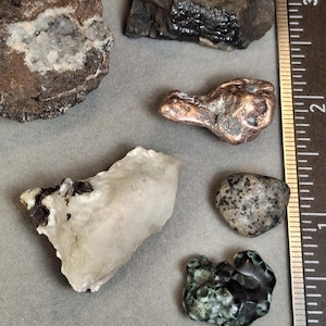 Può includere: Assortimento di varie rocce e minerali, tra cui una geode con formazioni cristalline, una pepita di rame e altri campioni. Un righello è incluso per la scala, con misure in centimetri.