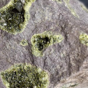 May include: Close-up of a rock formation with several pockets of bright green crystals. The rock has a rough, gray surface with a few small, clear crystals scattered throughout the green clusters. The overall texture is coarse and natural.