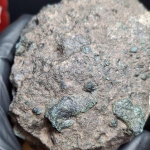May include: A rough, irregularly shaped rock with a dark brown and gray surface. Patches of dark green minerals are embedded within the rock, creating a textured appearance. The rock is held by a gloved hand.