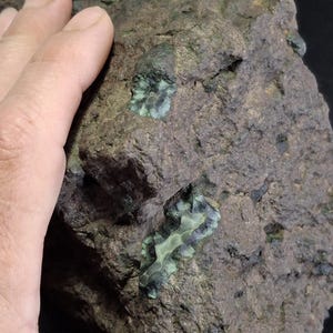 May include: A rough, brown rock with embedded, iridescent green and blue mineral formations. The formations have a layered, almost floral appearance. The rock's surface is textured and uneven, with a hand visible in the image for scale.