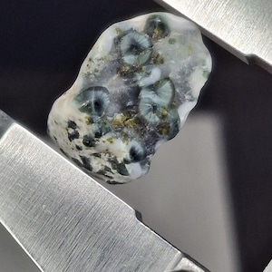 Puede incluir: Una piedra natural pulida con un patrón único de inclusiones verdes, blancas y doradas. La piedra tiene una forma suave y redondeada y está colocada sobre un fondo metálico. La apariencia general sugiere una gema o roca decorativa.