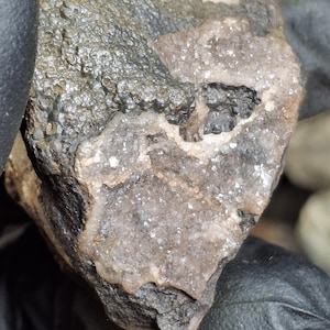May include: A close-up of a natural rock formation with a textured, dark gray surface. The rock has a crystalline interior with shades of gray and tan. The overall appearance suggests a geological specimen.