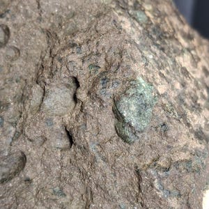 May include: Close-up of a rough, textured rock surface. The rock is primarily brown with areas of green and dark gray. The surface has small holes and irregular shapes, suggesting a natural, weathered appearance.