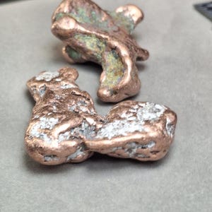 May include: Two irregularly shaped copper nuggets with a mix of brown, green, and silver tones. The nuggets have a rough, textured surface and are displayed against a gray background.