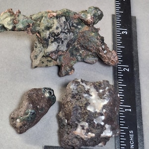 May include: Three irregularly shaped mineral specimens. The largest piece is approximately 3 inches long. The minerals display a mix of colors, including green, brown, and copper. A ruler is placed next to the minerals for scale.