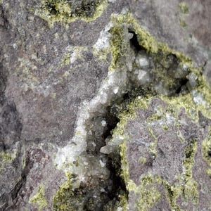 May include: Close-up of a rough, gray rock formation with a cavity filled with white crystals. The rock's surface is textured with patches of bright green mineral deposits, creating a striking contrast. The image highlights the natural beauty of the stone.