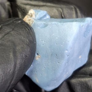 Puede incluir: Primer plano de una piedra azul claro de forma irregular. La piedra tiene una superficie rugosa y texturizada con pequeños agujeros y algunas zonas de color más claro. Un guante negro es parcialmente visible en el encuadre.