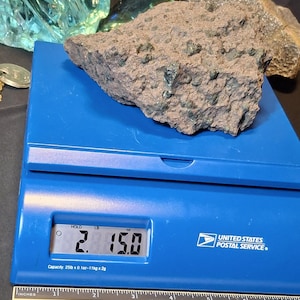 May include: A rough, reddish-brown rock with green inclusions sits on a blue scale. The scale displays a weight of 2.15 pounds. A ruler is visible below the scale, and the United States Postal Service logo is on the right.