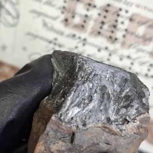May include: A close-up of a dark gray mineral specimen with a crystalline structure. The mineral has a metallic sheen and is set against a blurred background of text. The specimen is held by a gloved hand.