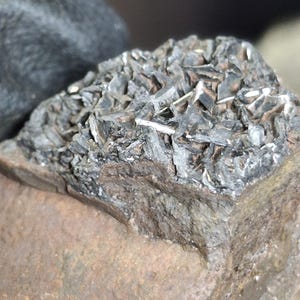 May include: Close-up of a mineral specimen featuring a dark, crystalline structure atop a reddish-brown rock base. The crystals have a metallic sheen, with sharp, angular forms. The rock's surface shows a rough, textured appearance.