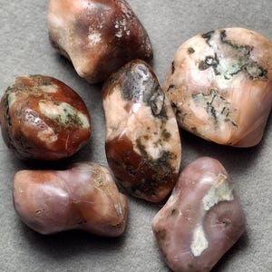 May include: A collection of six polished, tumbled stones in shades of dusty rose, terracotta, and cream. Each stone features a smooth, glossy surface with irregular shapes and natural inclusions, displaying mottled patterns of brown, white, and dark green.