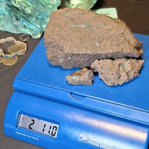 May include: A blue postal scale displays a weight of 2.1 pounds. Several pieces of rough, brown rock with green flecks are on the scale. Other gemstones and jewelry are in the background.