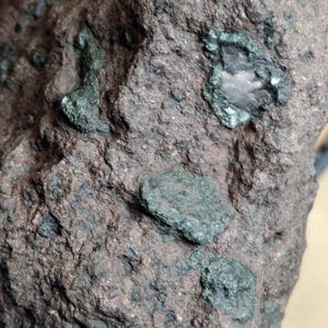 May include: Close-up of a rough, dark brown rock with embedded green mineral deposits. The rock's surface is textured and uneven, with various sizes and shapes of green inclusions scattered throughout. The overall appearance suggests a natural, geological specimen.