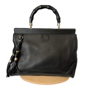 May include: Black leather handbag with a bamboo handle and gold-tone hardware. The structured bag features a top handle, a rectangular shape, and a tassel detail. The bag is sitting on a wooden surface.