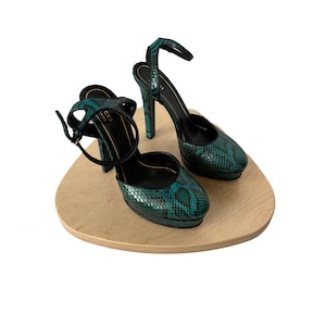 May include: A pair of high-heeled platform shoes with an ankle strap. The shoes feature a snakeskin pattern in shades of teal and black. The shoes are displayed on a light brown, oval-shaped platform.