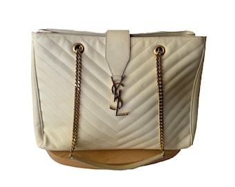 May include: Cream-colored leather tote bag with a chevron pattern. Features gold-tone chain straps and a gold logo. The bag has a flap closure and a rectangular shape. A stylish and elegant accessory.