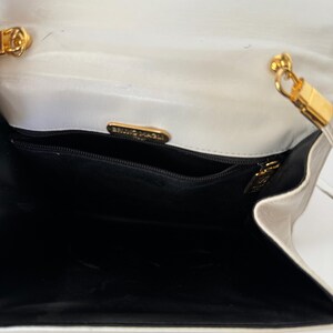 Puede incluir: Vista abierta de un bolso blanco con interior negro. El bolso presenta herrajes dorados, incluyendo una correa de cadena y un cierre. El nombre de la marca "BRUNO MAGLI" es visible en una placa dorada dentro del bolso.