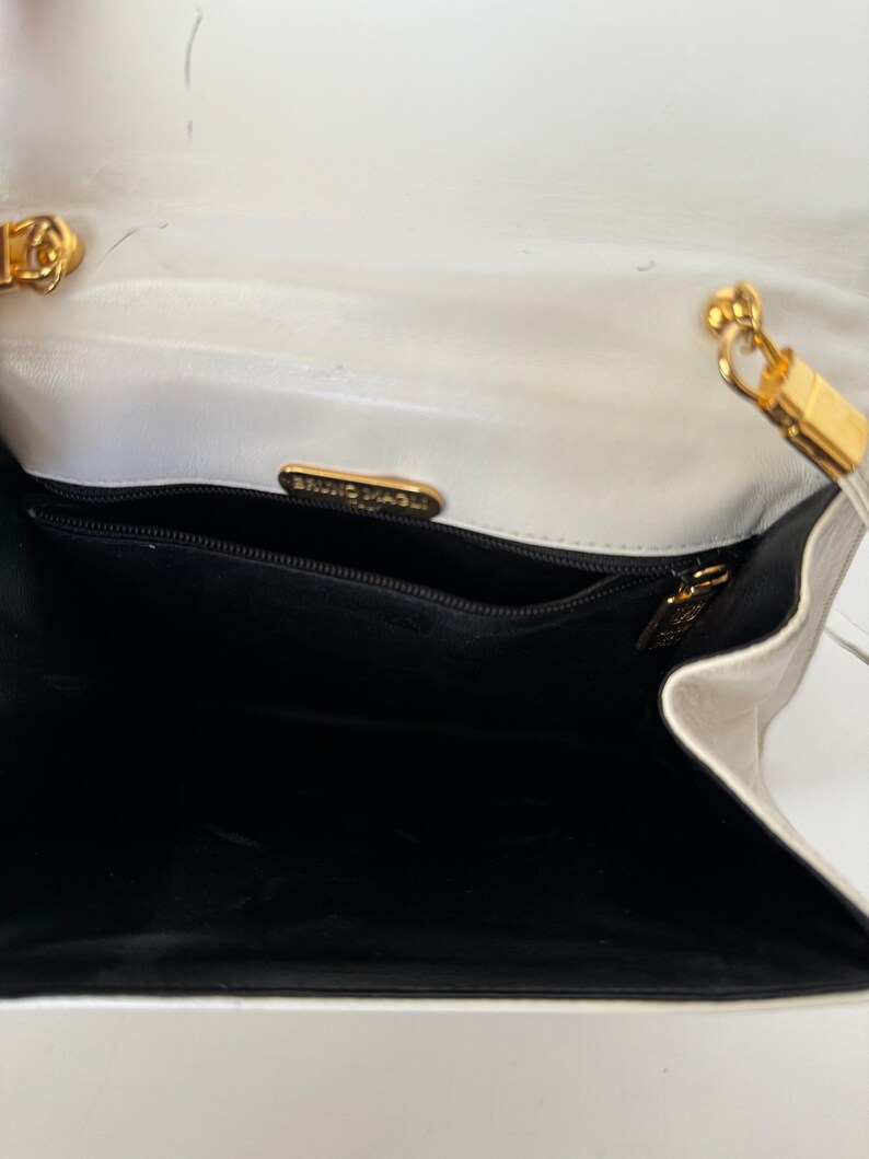 Puede incluir: Vista abierta de un bolso blanco con interior negro. El bolso presenta herrajes dorados, incluyendo una correa de cadena y un cierre. El nombre de la marca "BRUNO MAGLI" es visible en una placa dorada dentro del bolso.